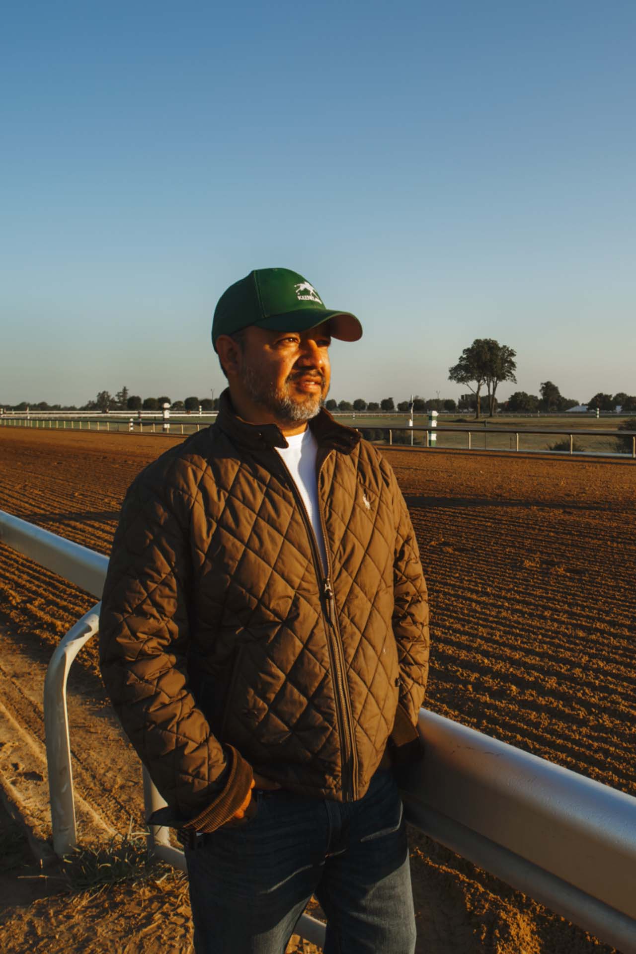 A photo of Alfredo Laureano looking off into the distance while standing by the dirt track at sunset. He is an older Hispanic man with graying chin stubble. He is wearing a green Keeneland-brand hat and a brown Keeneland-brand jacket overtop a white shirt.