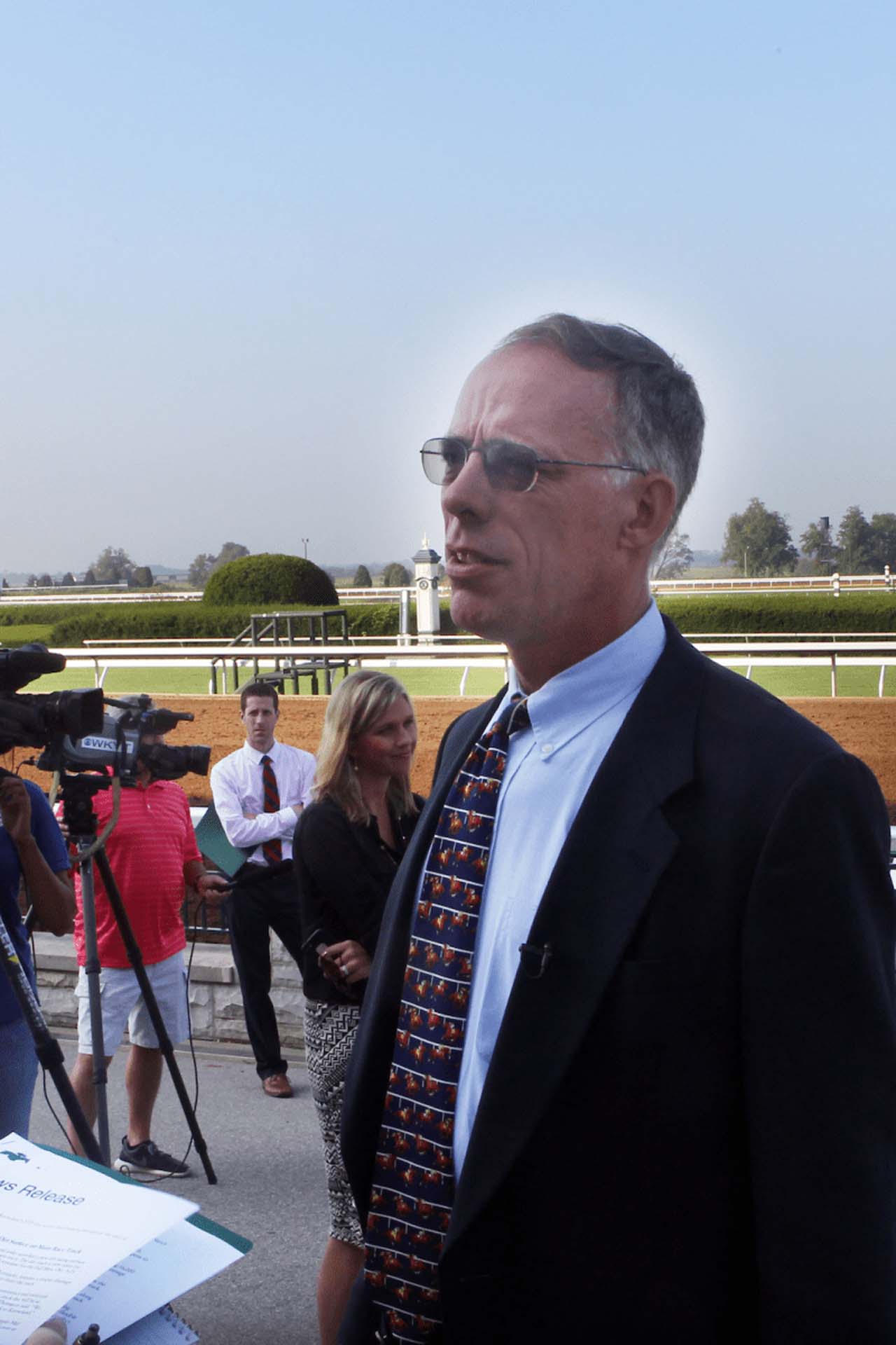 A candid photo of Dr. Mick Peterson giving an interview to an off-screen camera crew. He is an older white man with short gray hair. He is wearing a navy suit jacket over a light blue button-up shirt and a patterned tie. He is also wearing a pair of glasses with color-changing lenses.