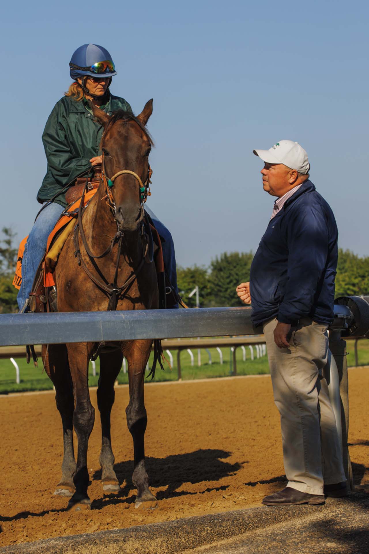 A candid photo of Dr. Stuart Brown leaning on a rail while talking to a woman who is riding a horse on the track. He is an older White man wearing a white hat, a navy pullover sweater, and khakis.
