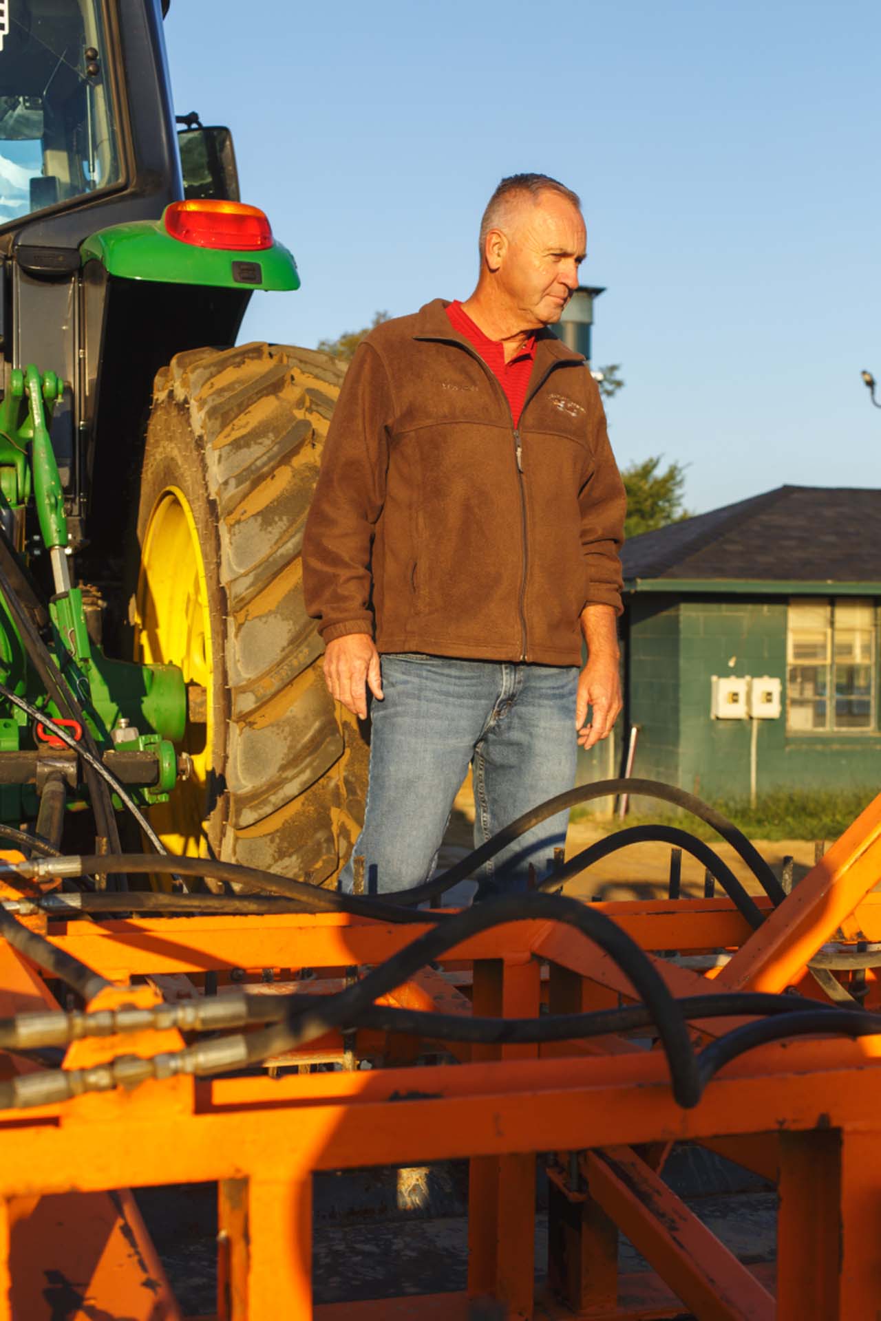 A candid photo of Jim Pendergest standing in front of a large piece of equipment. He is an older White man with short gray hair. He is wearing a brown jacket over a red polo and a pair of blue jeans.