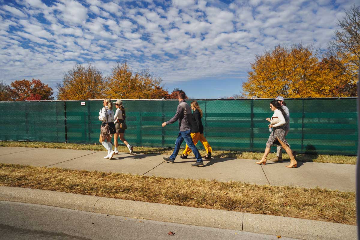 A photo of six people walking on a sidewalk in front of a green fence