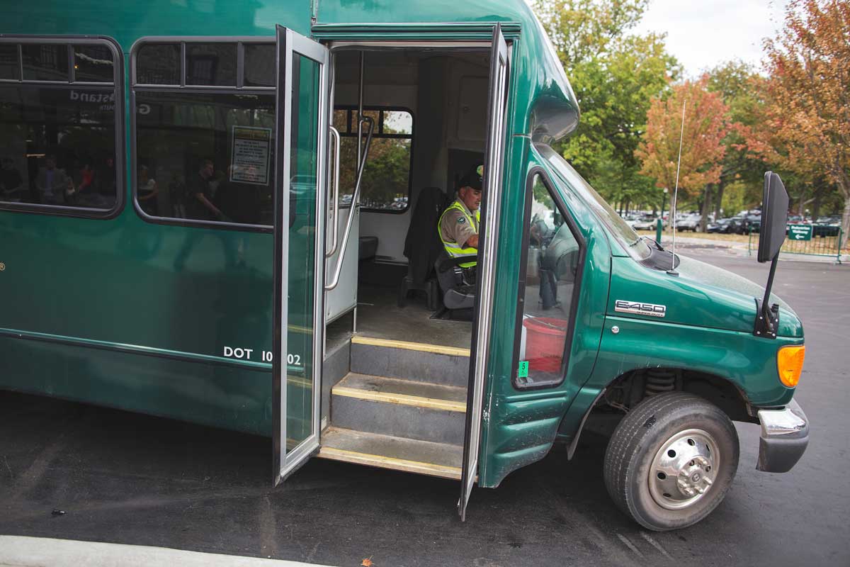Photo of a green shuttle bus with an open door. There is a man in the driver's seat wearing a high visibility vest.